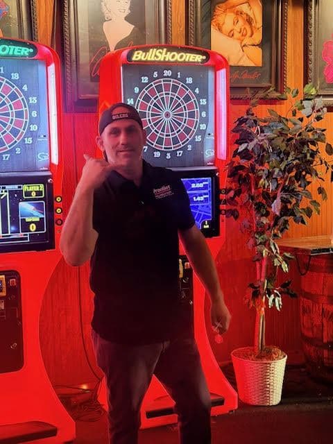 A regular posing in front of two BullShooter electronic dart machines under red light, framed pin-up prints on the wall beside them.