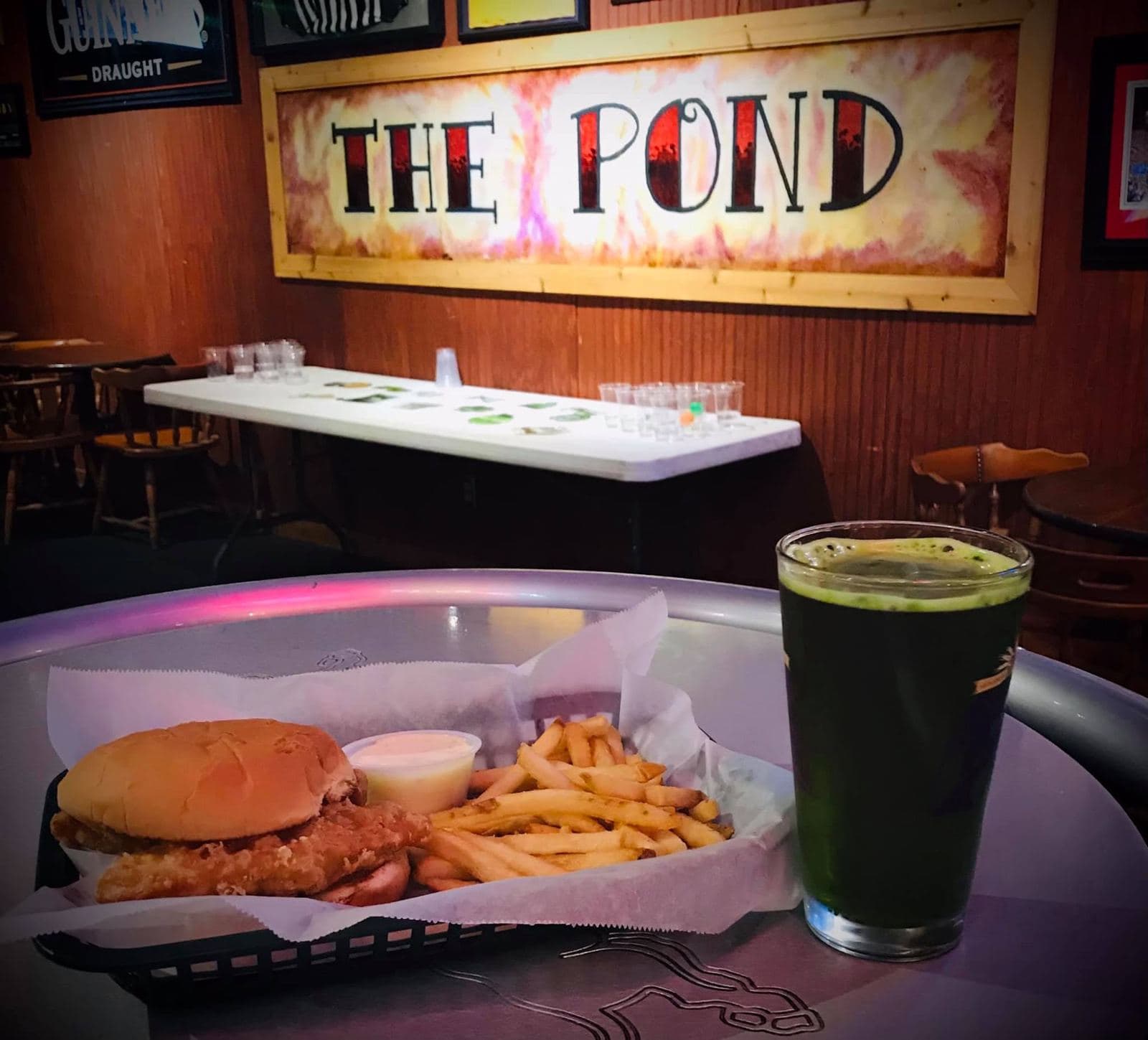 A hand-painted THE POND sign on the panelled wall, with a chicken sandwich and fries in a basket and a dark pour on the table in front of it.