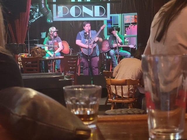 A four-piece band mid-song, seen past two glasses on a table at the back of the room.