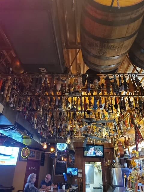Tap handles hanging in racks beneath a mounted whiskey barrel, bartenders serving guests at the bar below.