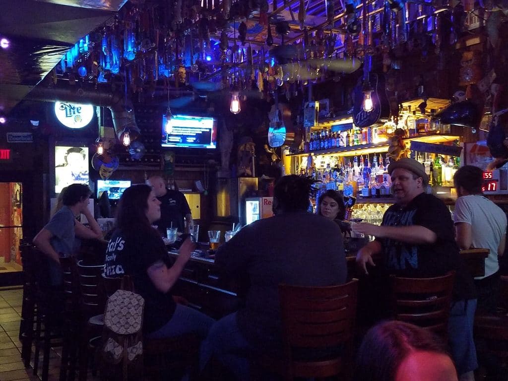 Drinkers on stools along the bar in the early evening, blue-lit tap handles above and the back-bar bottles glowing behind.