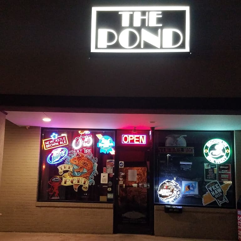 The Pond's storefront at night: a lit box sign reading THE POND above windows filled with neon beer signs and a red OPEN sign by the door.