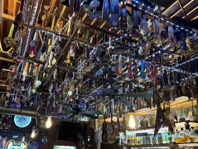 A ceiling densely packed with rows of beer tap handles, string lights threaded between them and two ceiling fans hanging below.