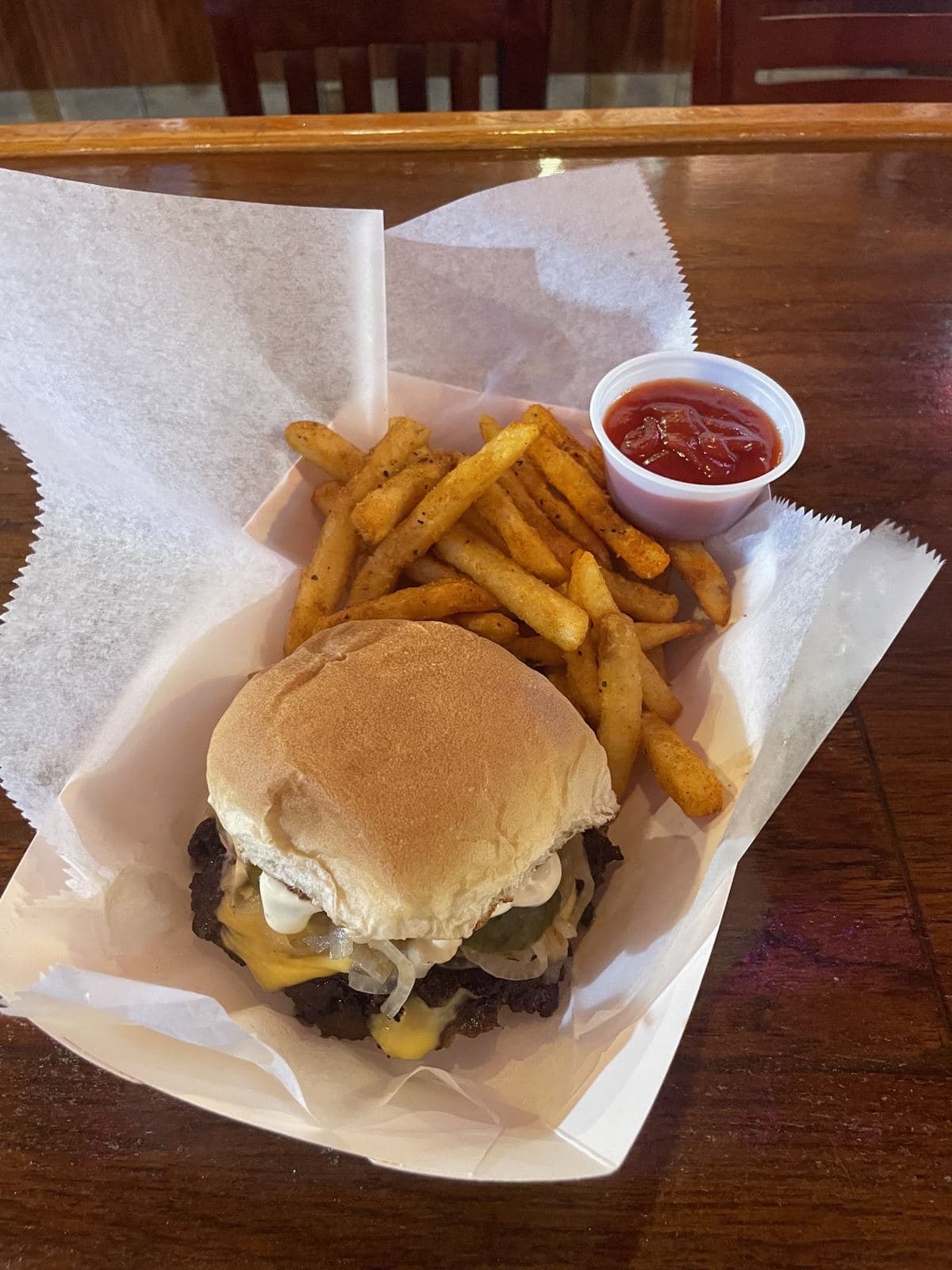 A cheeseburger with grilled onions and pickles on a toasted bun, in a paper-lined basket with seasoned fries and a cup of ketchup.