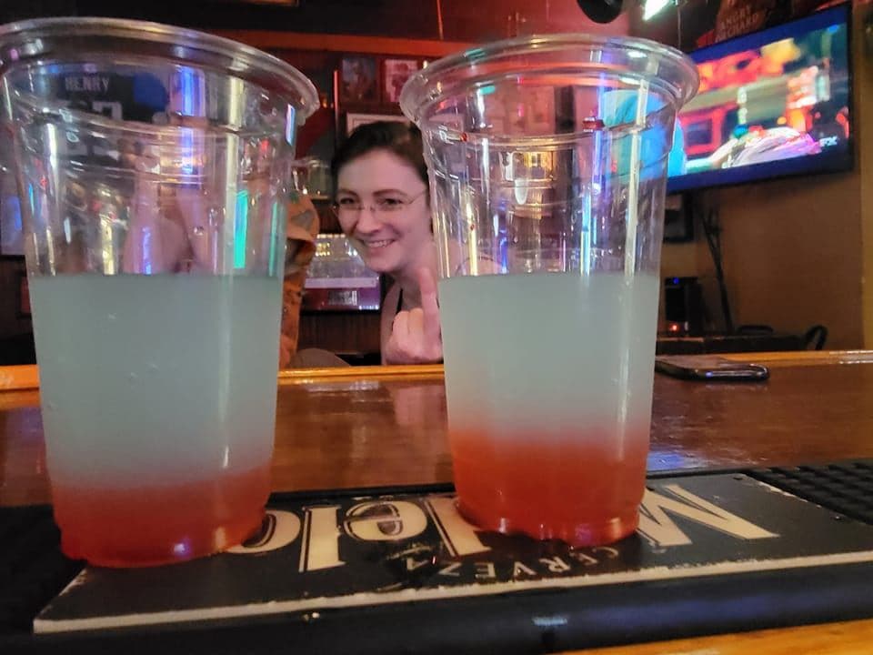 Two layered red-and-clear cocktails in cups on the bar, the bartender smiling behind them.