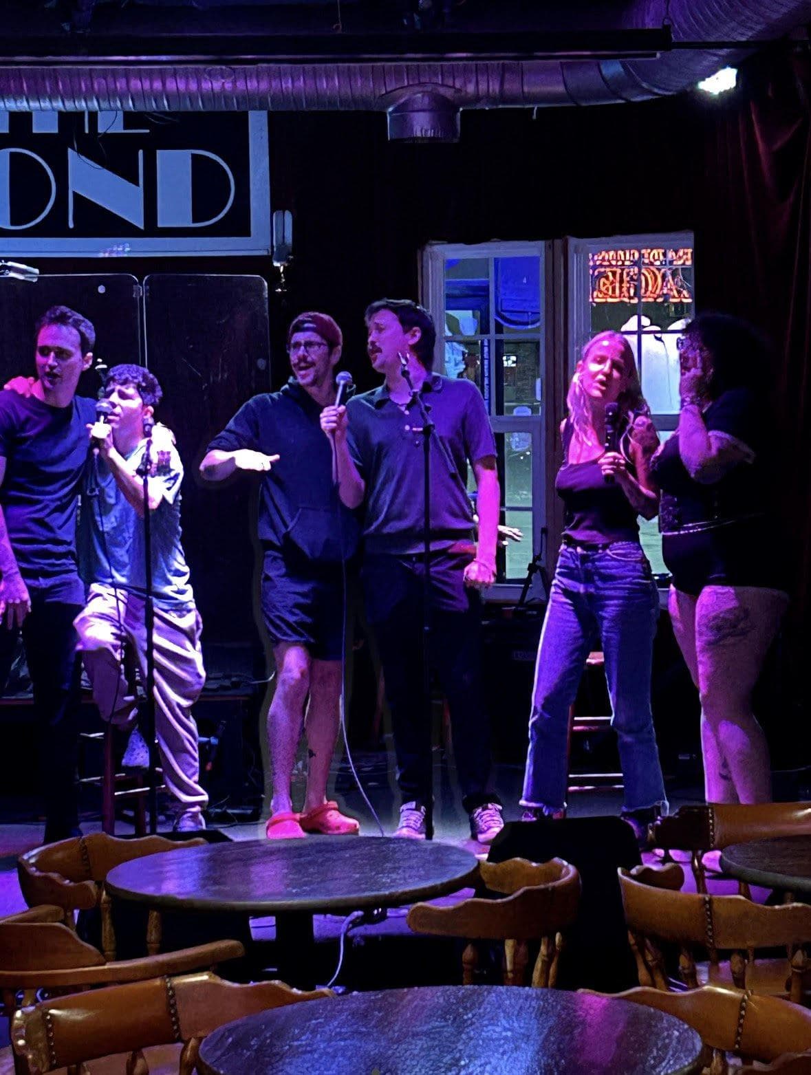Six people crowded onto the stage sharing microphones under purple light, the tables in front of them empty.