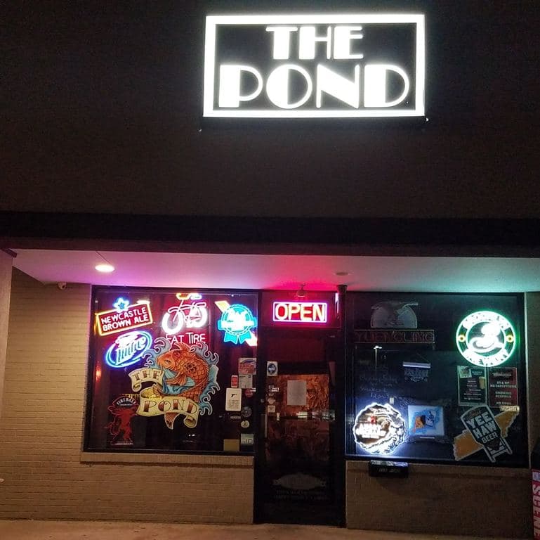 The storefront at night: a lit box sign reading THE POND above windows full of neon beer signs, a red OPEN sign and the koi emblem in the glass.