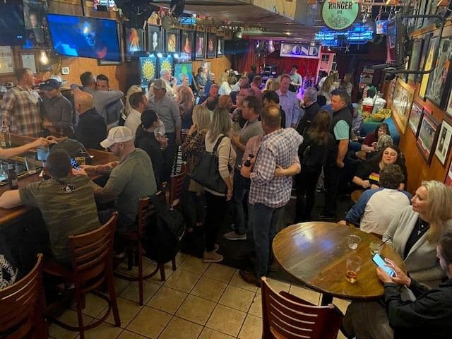 A busy night with the room full, people standing shoulder to shoulder between the bar and the tables and a band playing at the far end.