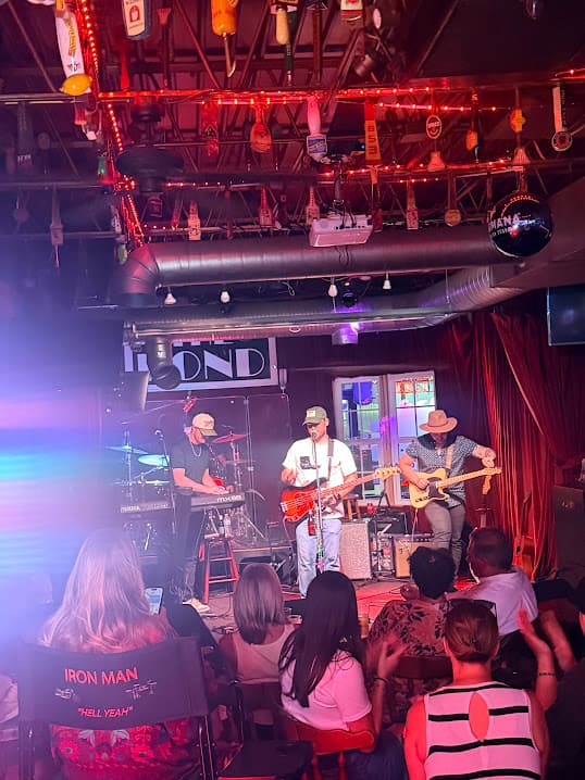 A three-piece band on stage under blue and pink light, tap handles strung with lights across the ceiling above, a seated crowd watching in front of them.