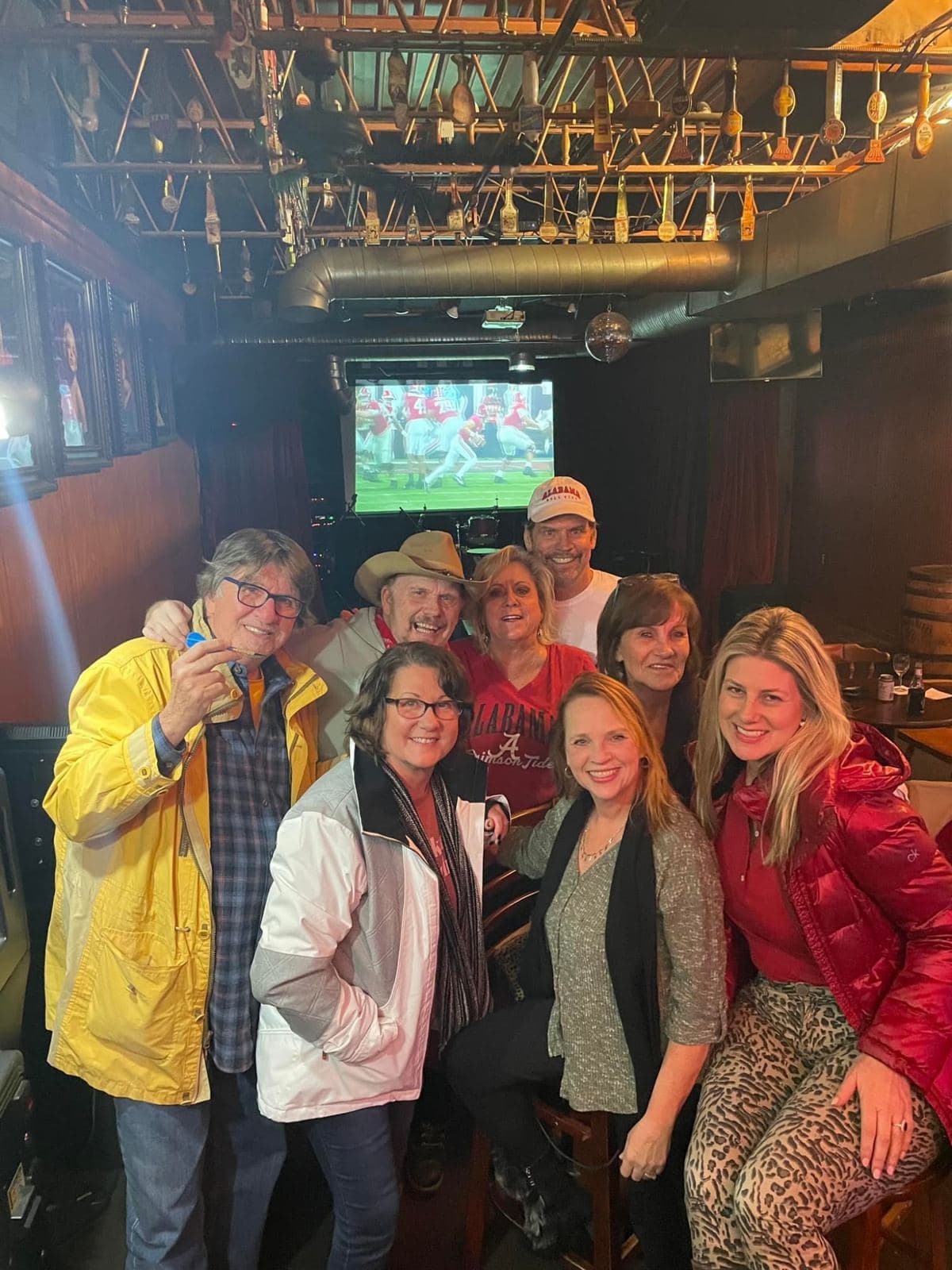 Eight guests packed together for a photograph under the tap-handle ceiling, a football game on the screen behind them.