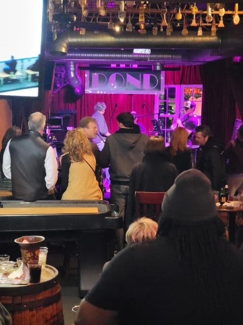 The stage seen over the heads of the crowd, a band playing under pink light with tap handles hanging from the ceiling above.